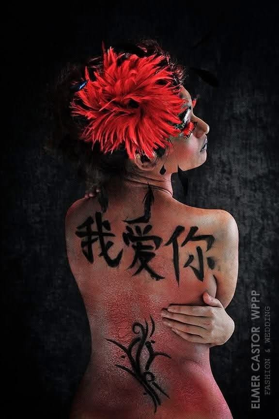 Model photographed from behind with bold red body paint and calligraphic characters across her back, wearing a red feather headpiece.
